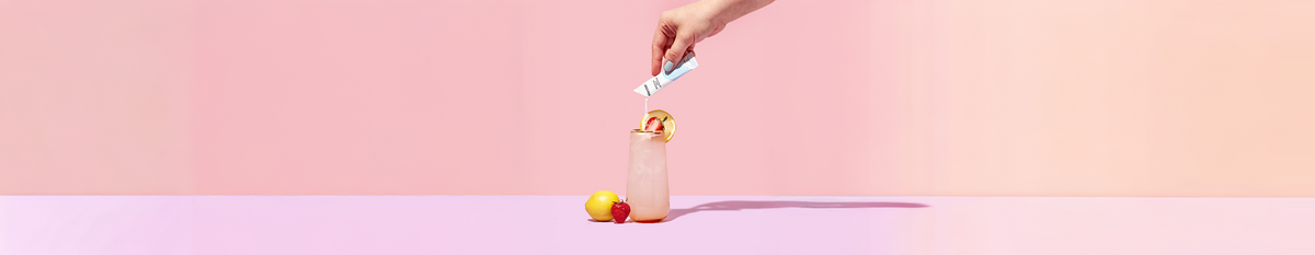Hand pouring powdered drink mix into a tall ice-filled glass garnished with lemon and strawberry on a pink gradient background